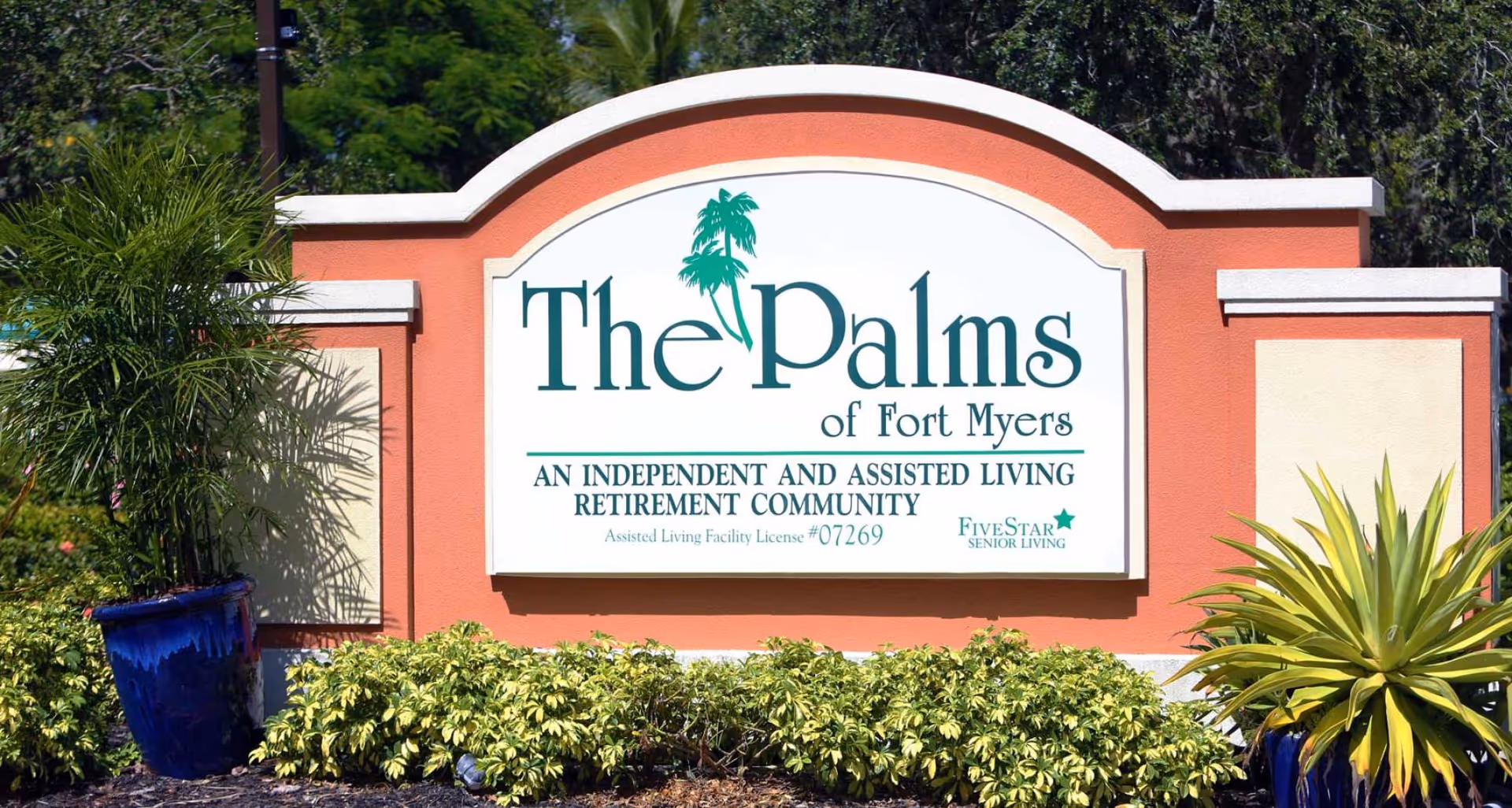 Entrance sign for The Palms of Fort Myers, an independent and assisted living retirement community, surrounded by green plants and trees.
