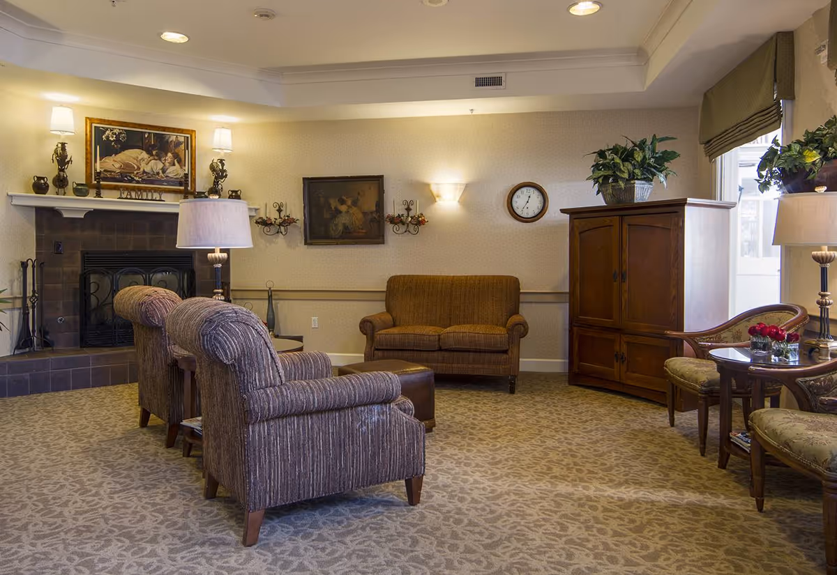 A cozy living room with patterned carpet, two upholstered armchairs facing a fireplace with a painting above it, a brown loveseat against the wall, a wooden cabinet with plants on top, and a small round table with two chairs and a lamp near a window.