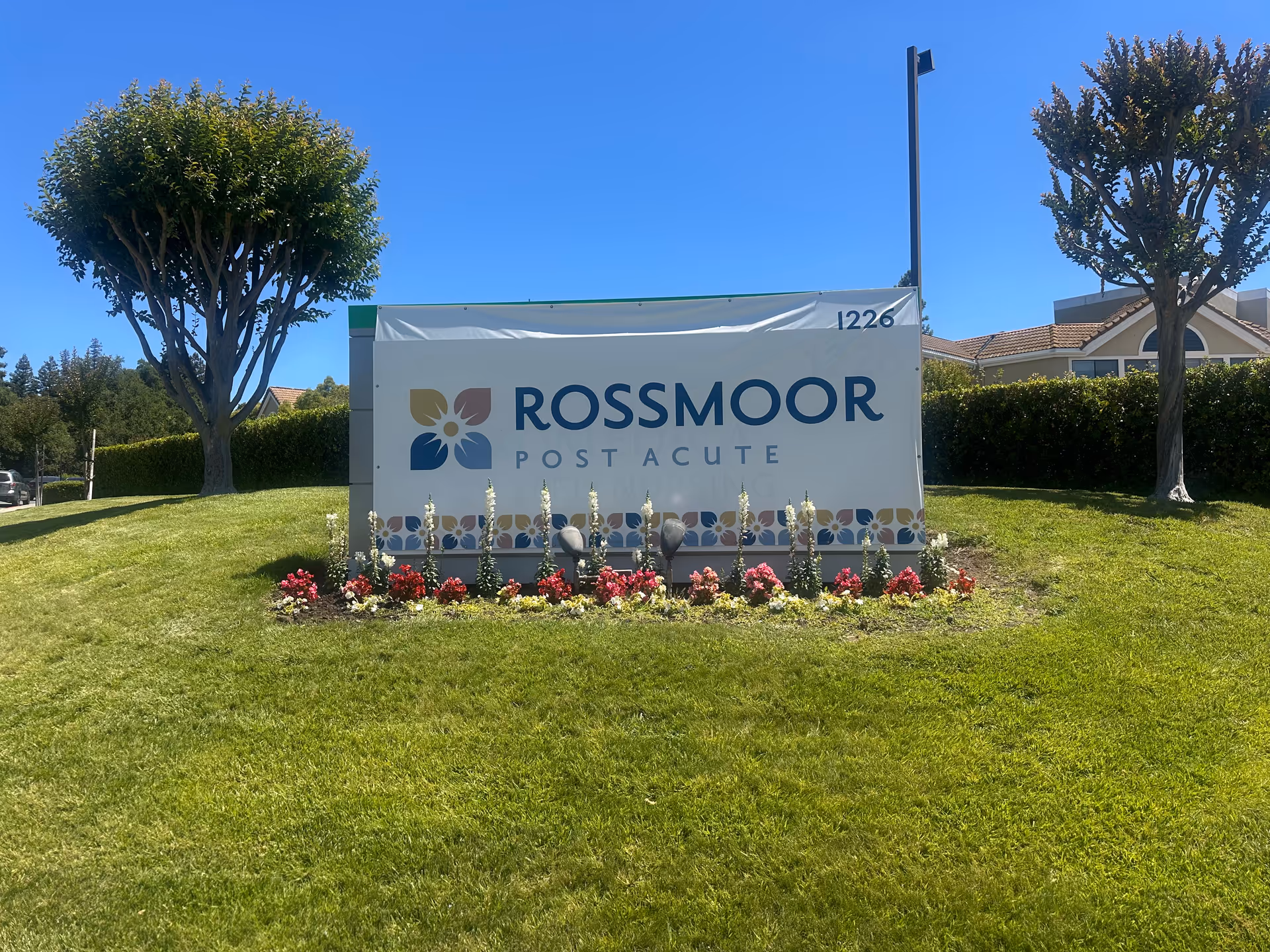 Outdoor landscaped sign reading 'Rossmoor Post Acute' on a grassy lawn with flowers and trees under a clear blue sky.
