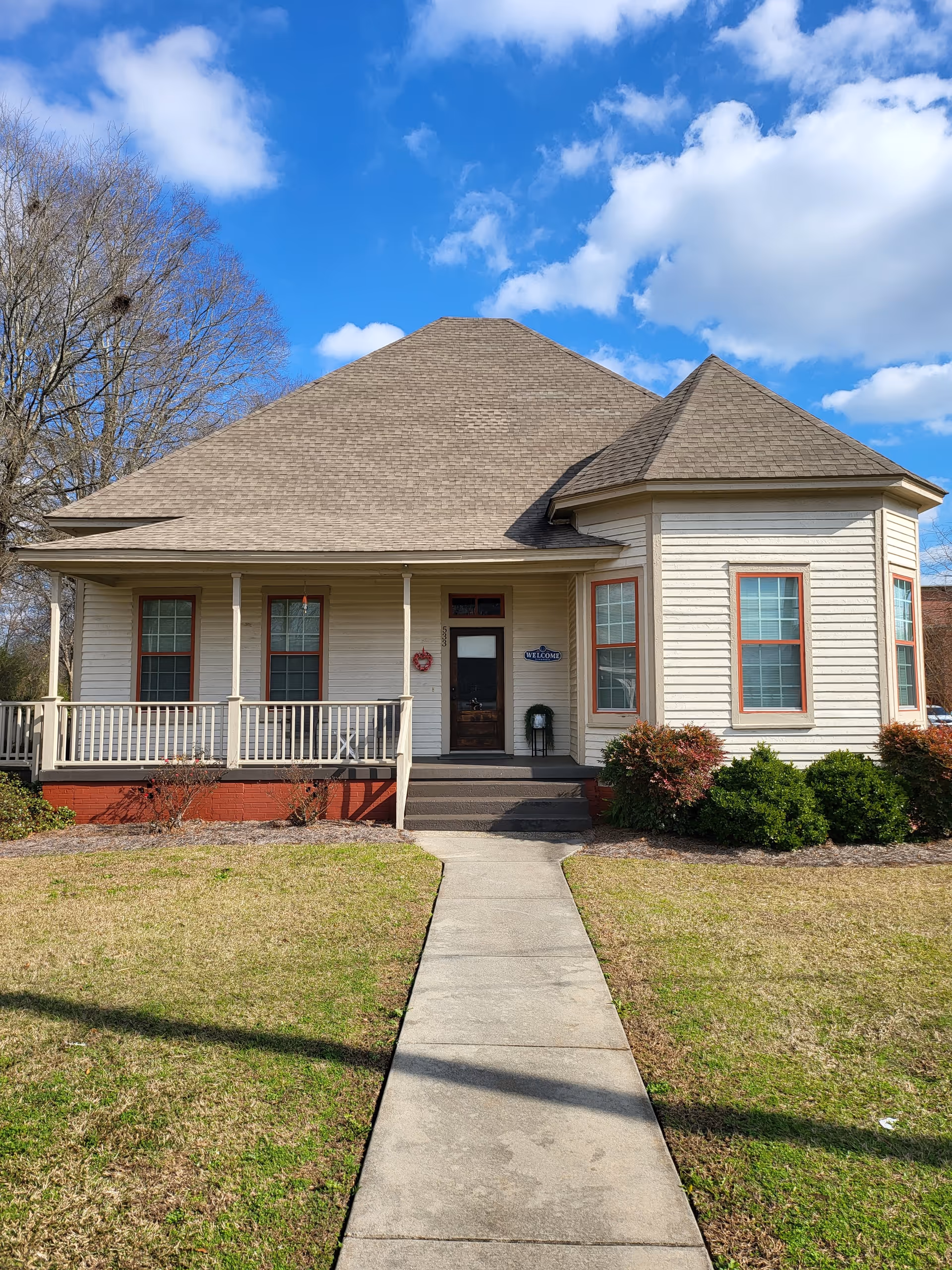 Front exterior view of a single-story house with beige siding, a brown shingled roof, and a small porch with white railings. There is a concrete walkway leading up to the front door, which has a wreath and a welcome sign beside it. The house is surrounded by a lawn with some bushes and trees, under a partly cloudy blue sky.