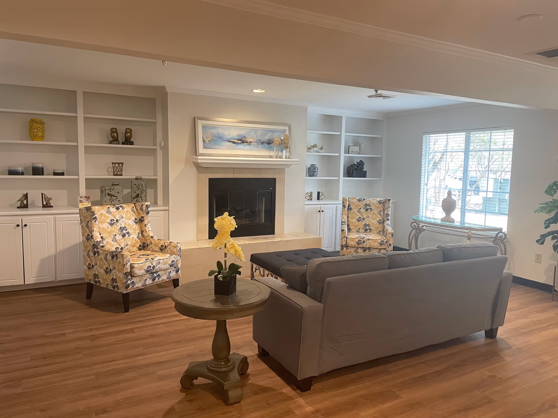 A cozy living room area with a gray sofa, two floral-patterned armchairs, a round wooden side table with a yellow orchid plant, built-in white shelves with decorative items, a fireplace with a painting above it, and a window with blinds letting in natural light.