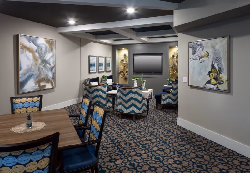 A cozy interior common area with patterned carpet and walls adorned with abstract and bird paintings. There are several upholstered chairs with blue and beige zigzag patterns arranged around a small table, and a flat-screen TV mounted on the far wall. A wooden table with matching chairs is visible in the foreground.
