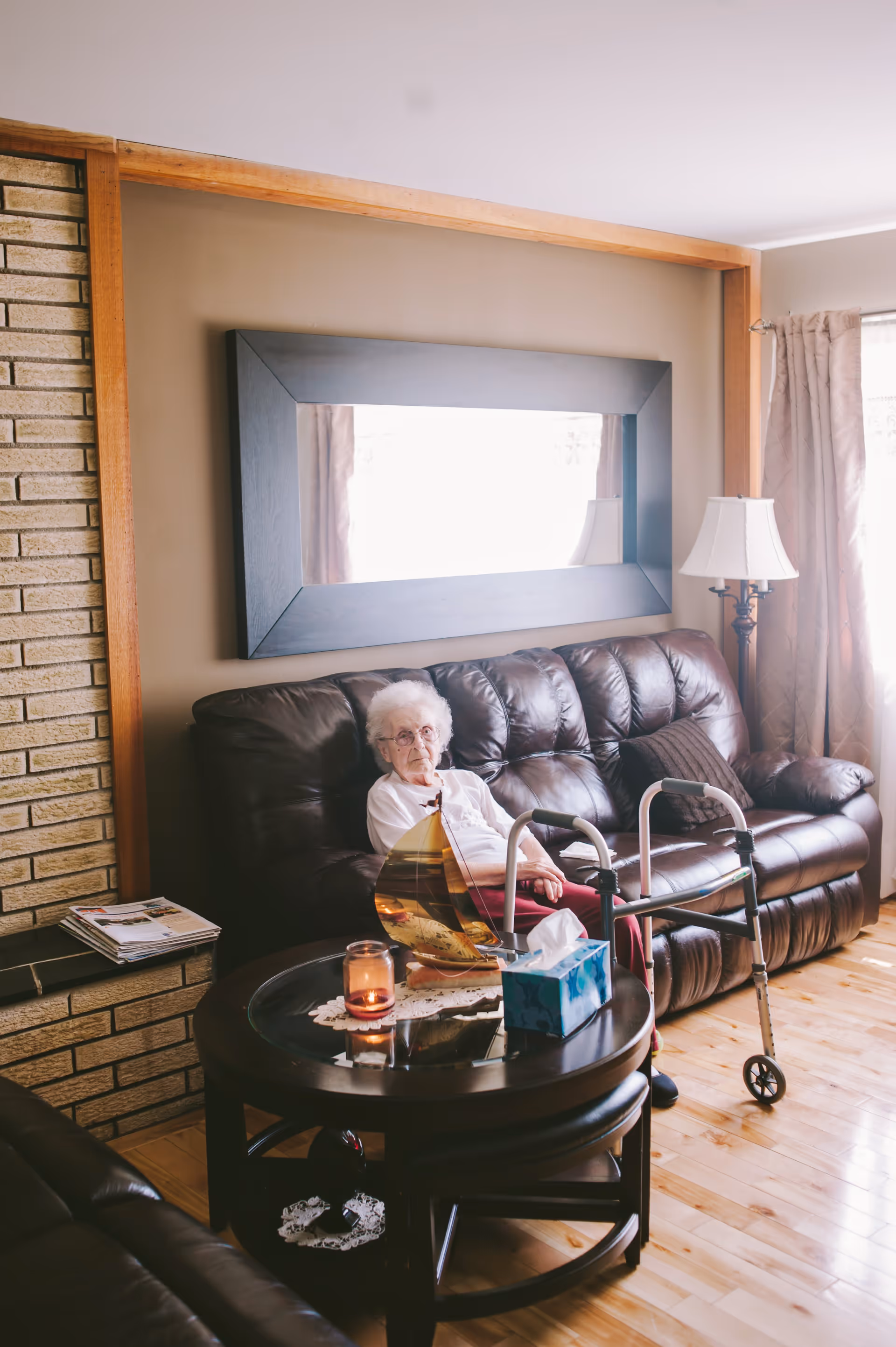 An elderly woman sitting on a dark brown leather couch in a living room. She has white hair and glasses and is wearing a white top and red pants. In front of her is a round wooden coffee table with a candle, a tissue box, and a decorative item. A walker is positioned next to the couch. The room has wooden flooring, a large rectangular mirror on the wall, a floor lamp, and a window with curtains letting in natural light.