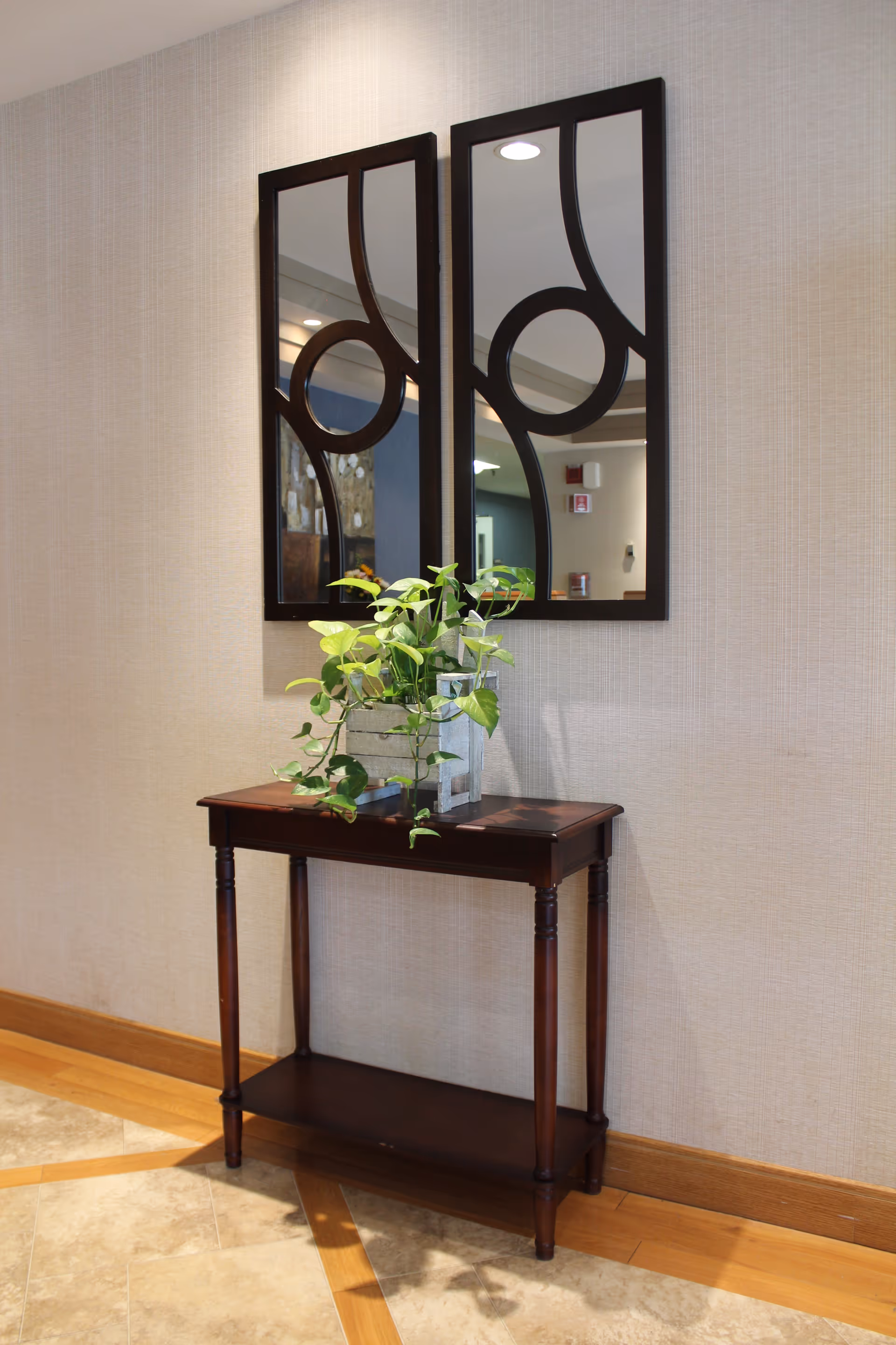 A small wooden console table with a potted green plant on top, placed against a beige textured wall. Above the table are two rectangular mirrors with black frames and decorative circular patterns. The floor is tiled with a combination of light and medium brown tiles.