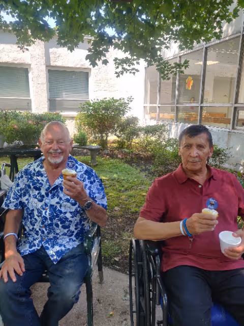 Two elderly men sitting outside in a garden area under a tree. One man is wearing a blue floral shirt and holding a small cup, smiling. The other man is wearing a red polo shirt, sitting in a wheelchair, also holding a small cup and a white bowl. Behind them are bushes, windows, and part of a building.