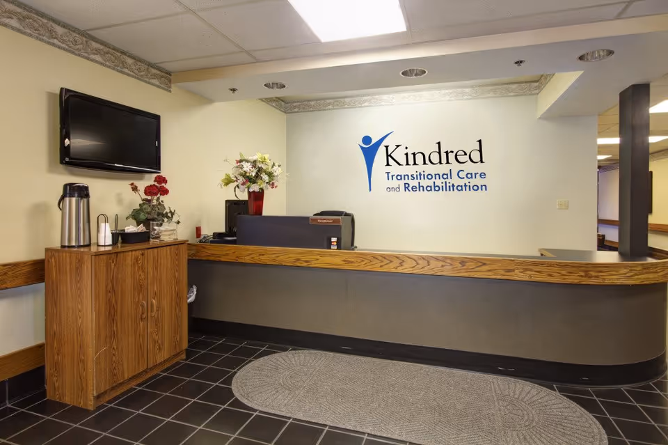 Reception desk area with a Kindred Transitional Care and Rehabilitation logo on the wall, a TV, flowers, and a small coffee station.