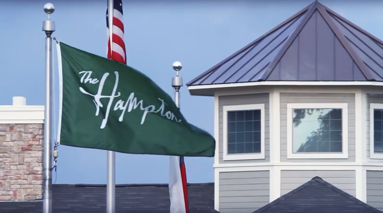 A green flag with the text 'The Hamptons' flying on a flagpole next to an American flag, with part of a building featuring a gray roof and windows in the background.