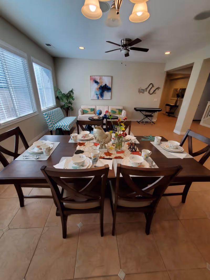 A dining area with a dark wooden table set for six people with plates, cups, and cutlery. The table has a decorative centerpiece with flowers and a rooster figurine. In the background, there is a living area with a white couch, a patterned chair, a small table with a globe, and a keyboard against the wall. The room has large windows with blinds, a ceiling fan, and tiled flooring.