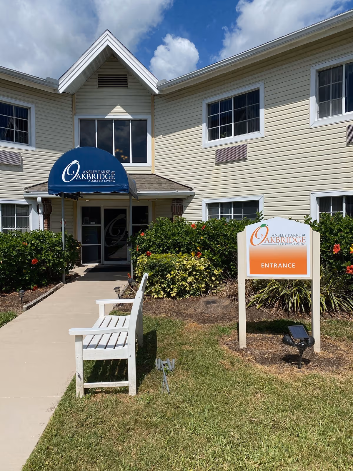 Front entrance of Ansley Parke at Oakbridge assisted living with a blue awning, entrance sign, walkway, and a white bench.
