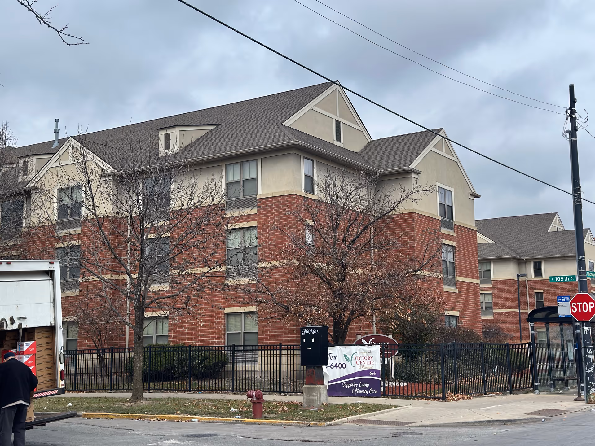 Exterior view of a multi-story brick-and-stucco senior living building with a fence and a Victory Centre sign at the corner.