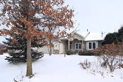 A single-story house with a snow-covered yard and trees with brown leaves in front. The house has a garage, a front porch, and multiple windows. The sky is clear and bright.