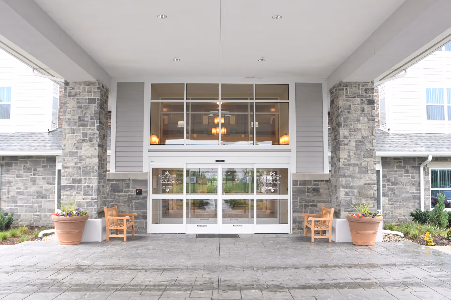 Entrance of Rolling Hills Retirement Community featuring automatic sliding glass doors framed by stone pillars, two wooden benches on either side, large potted plants with flowers, and a covered driveway.