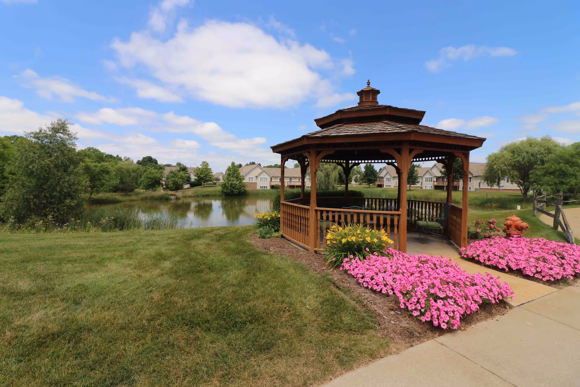 A wooden gazebo surrounded by vibrant pink and yellow flowers sits next to a small pond with green trees and residential buildings in the background under a partly cloudy blue sky.