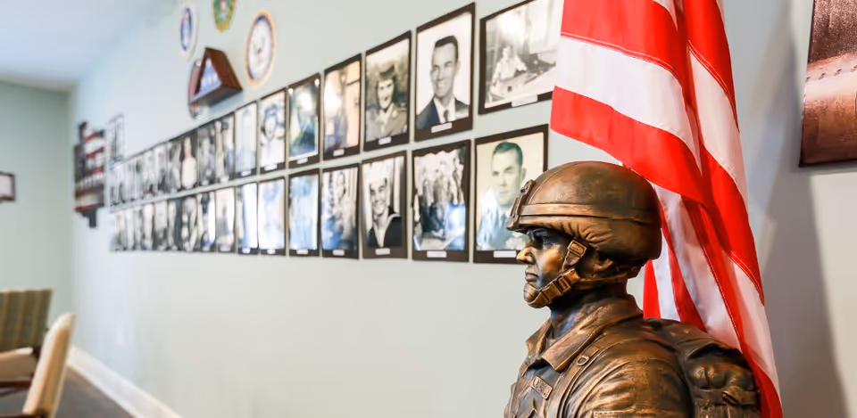 Bronze soldier statue beside an American flag in a corridor lined with framed black-and-white portraits on a pale green wall.
