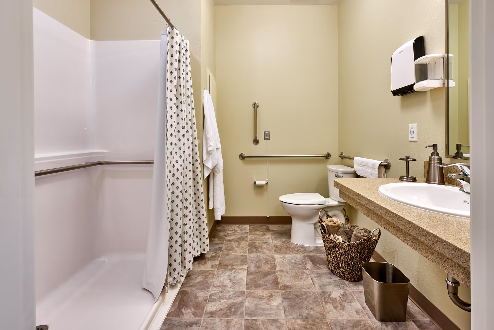 A clean and accessible bathroom with a walk-in shower featuring a patterned shower curtain, grab bars on the walls, a white toilet, a countertop sink with soap dispenser, a wall-mounted paper towel dispenser, a basket with rolled towels, and a small trash bin on a tiled floor.