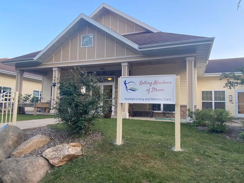 Front exterior view of a single-story building with beige siding and a gabled roof. There is a sign in front that reads 'Rolling Meadows of Strum Assisted Living and Memory Care'. The building has a covered porch area with columns and some greenery and rocks in the foreground.