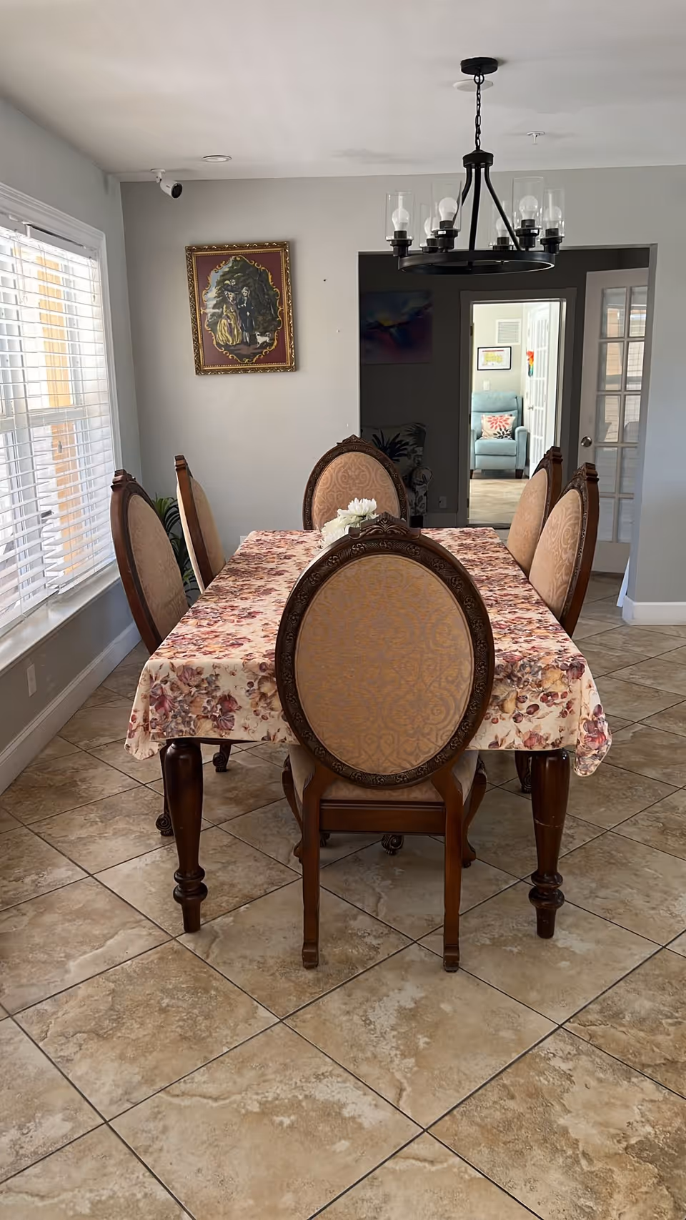 A dining room with a rectangular wooden table covered with a floral tablecloth and six upholstered chairs with ornate wooden backs. The room has beige tiled flooring, a window with white blinds on the left, a chandelier hanging above the table, and a framed painting on the wall. In the background, there is a doorway leading to another room with a light blue armchair and some wall decorations.