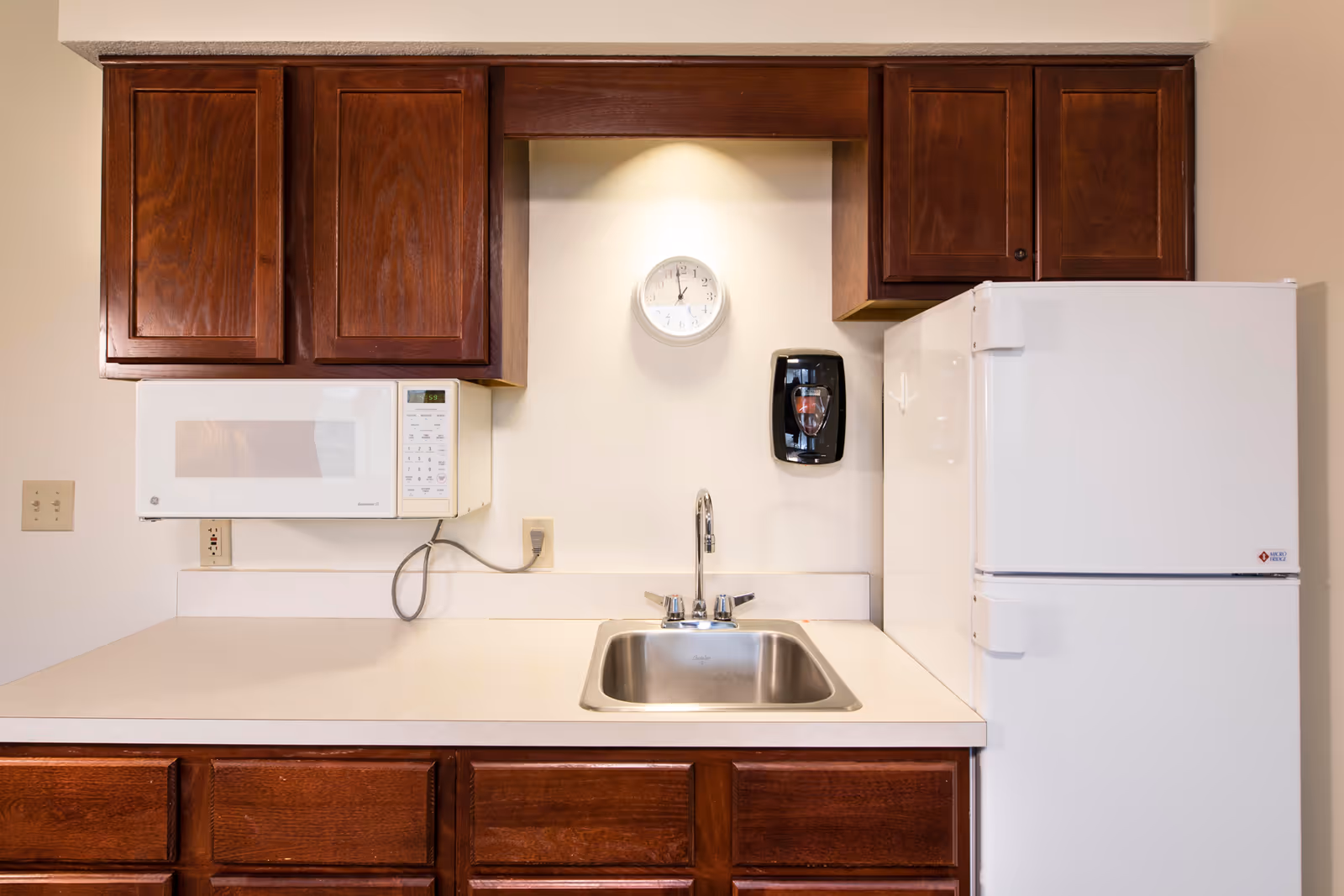 Small kitchenette with a stainless steel sink, microwave, white refrigerator, wooden cabinets, and a wall clock.