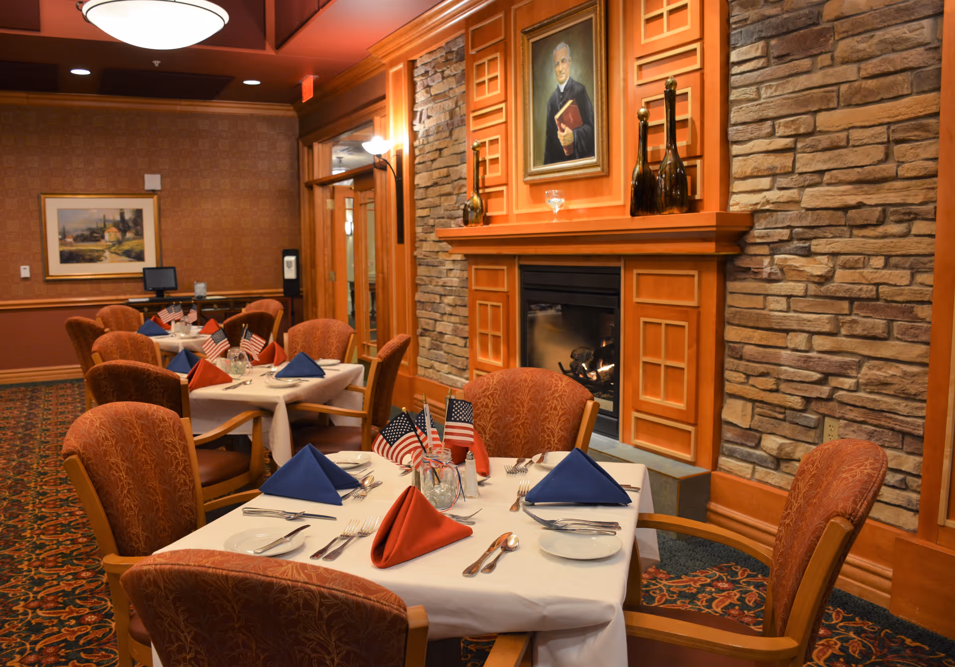A warmly lit dining room with tables set for a meal, featuring white tablecloths, folded red and blue napkins, and small American flags as centerpieces. The room has upholstered chairs, a stone fireplace with a portrait above it, and patterned carpet and wallpaper.