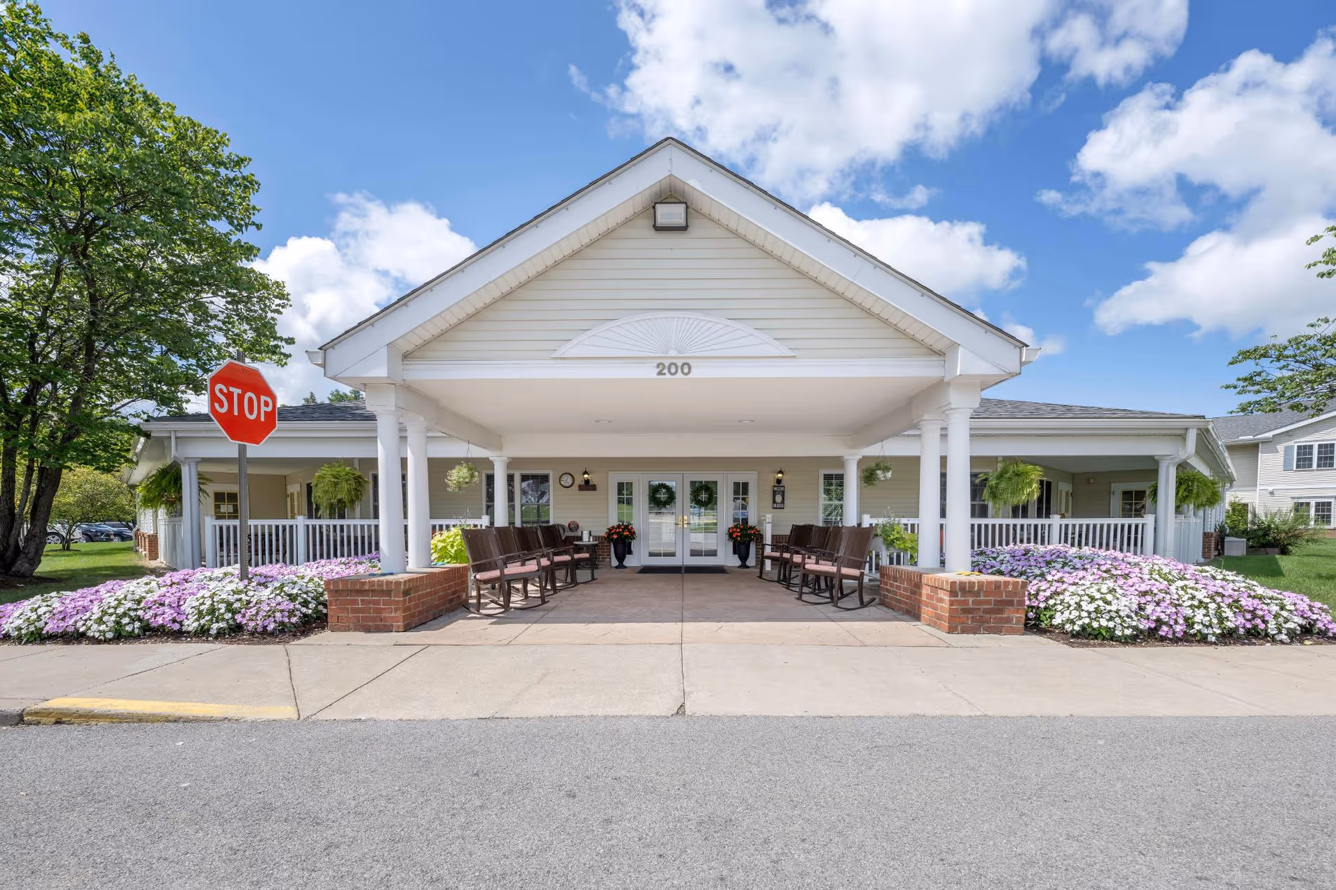 Front entrance of a senior living facility with a covered porch supported by white columns, benches on either side, flower beds with purple and white flowers, a stop sign on the left, and a clear blue sky with some clouds.