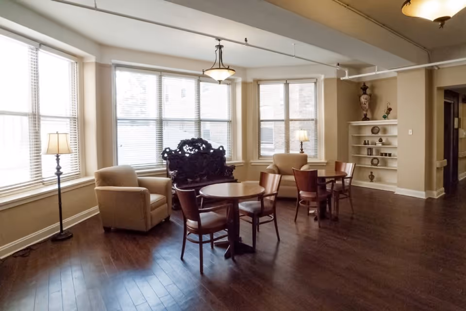 A well-lit common area with large windows covered by blinds, featuring two beige armchairs, two small round wooden tables each surrounded by wooden chairs, a decorative dark wooden bench, a floor lamp, and a built-in shelf with decorative items against a beige wall.
