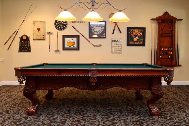 A classic wooden pool table with green felt in a game room. The wall behind the table is decorated with various game-related items including a dartboard, billiards-themed artwork, a bingo card, and several cues and racks for pool sticks. The room has patterned carpet and a three-light fixture hanging above the pool table.