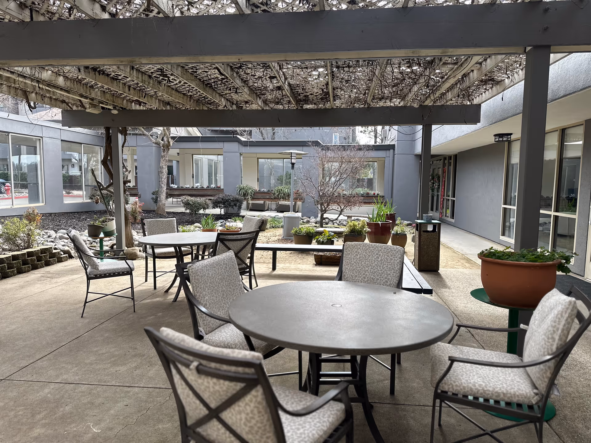 Outdoor patio area at Gold Country Senior Living with round tables and cushioned chairs under a pergola. There are potted plants and shrubs around the seating area, with the building's windows and doors visible in the background.