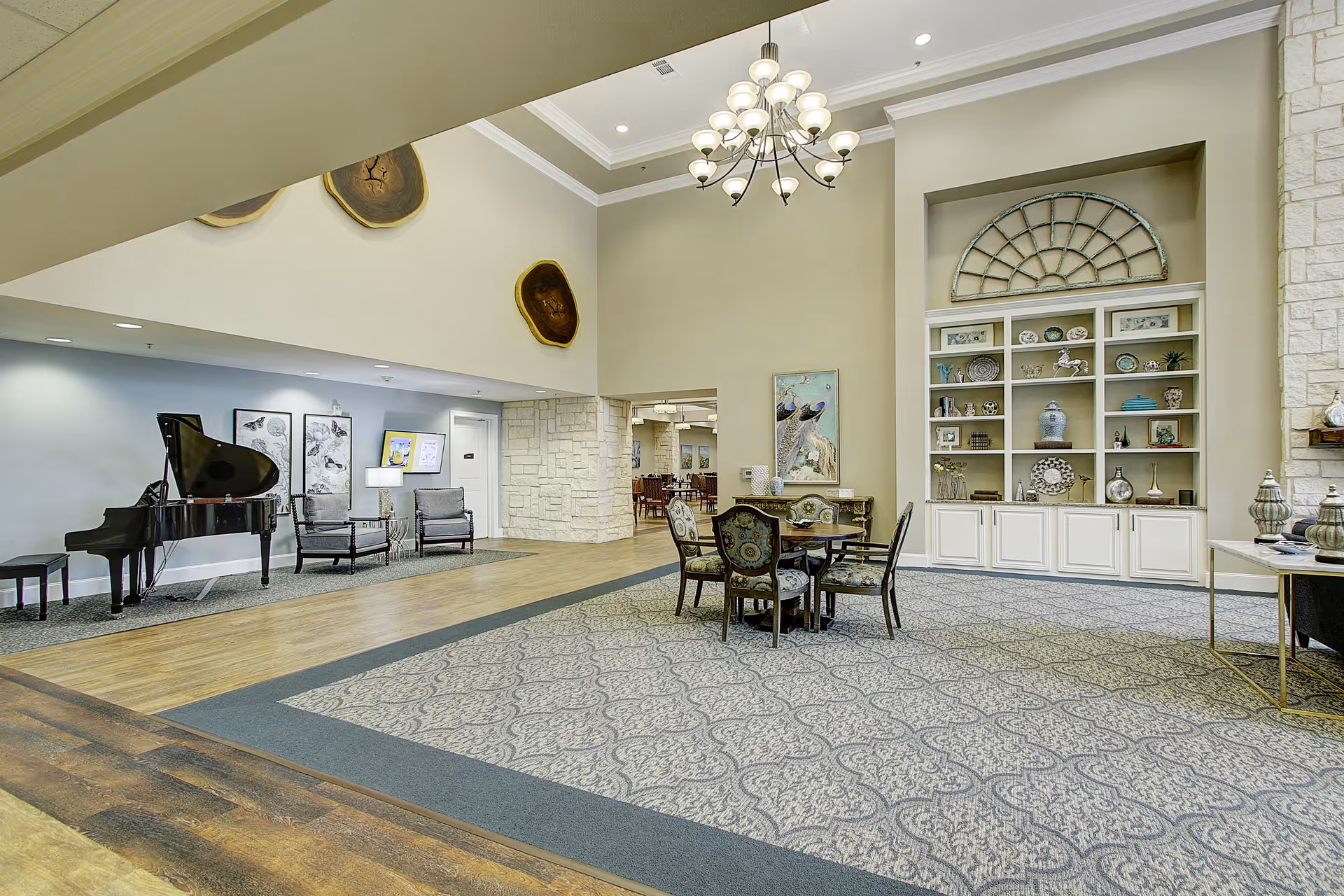 A spacious and well-lit common area with a grand piano on the left, two armchairs, a round table with four upholstered chairs in the center, and built-in shelves displaying decorative items on the right. The room features high ceilings with a chandelier, light-colored walls, and a patterned carpet.