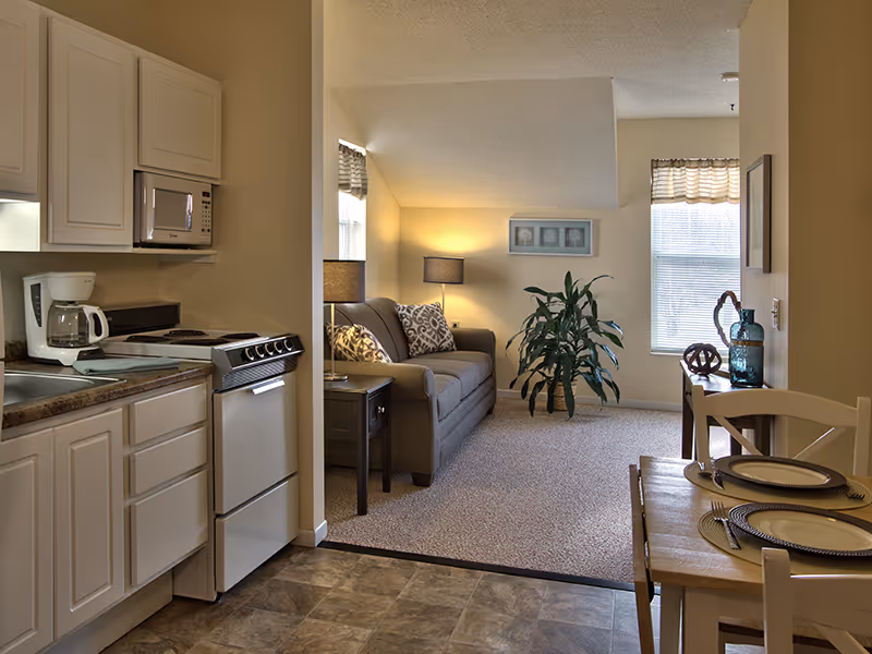 Interior view of a senior living facility apartment showing a small kitchen area with white cabinets, a stove, microwave, and coffee maker on the left. Adjacent to the kitchen is a living room with a gray sofa, side table with a lamp, a large green plant, and framed artwork on the wall. To the right is a dining area with a wooden table set with plates and placemats, and a window with blinds letting in natural light.