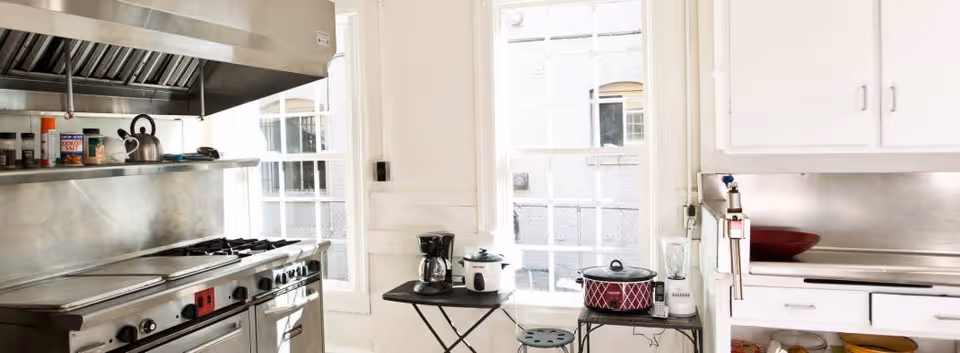 A bright kitchen with large windows letting in natural light. The kitchen features a stainless steel stove and oven with a vent hood above, a shelf with various spices and a kettle, and a small table holding a coffee maker, rice cooker, slow cooker, and blender. White cabinets and a stainless steel backsplash are also visible.