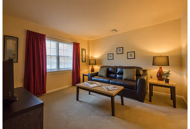 A cozy living room with a dark leather sofa adorned with two pillows, a wooden coffee table with magazines, two side tables each with a lamp and a small plant on one, framed artwork on the walls, a window with red curtains, and a television on a stand.