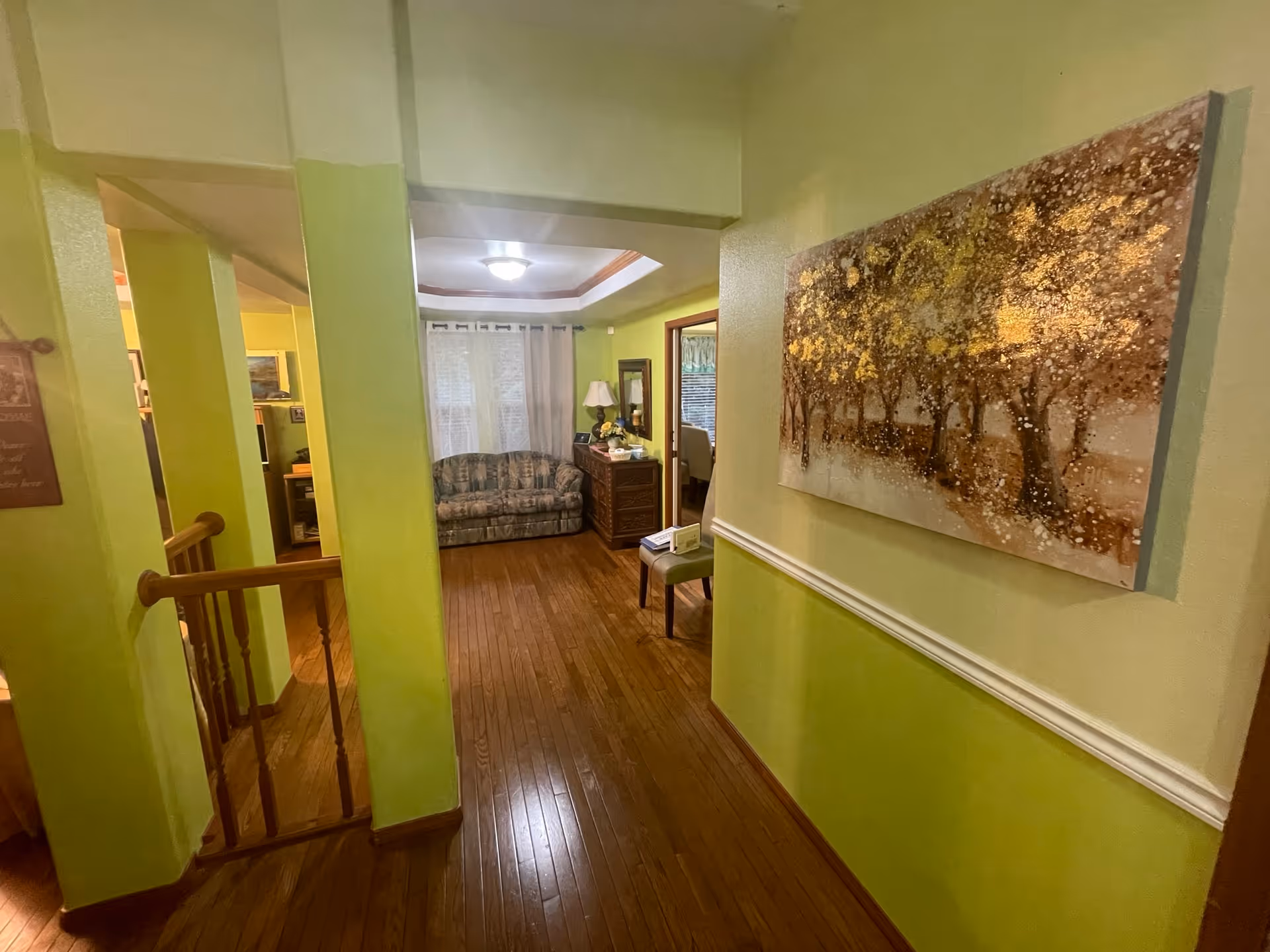 Interior view of a hallway in a senior living facility with light green walls and wooden flooring. On the right wall, there is a large painting of trees with golden and brown tones. At the end of the hallway, there is a small sitting area with a patterned couch, a wooden dresser with a lamp, and a window with white curtains. To the left, there is a staircase with wooden railings leading down.