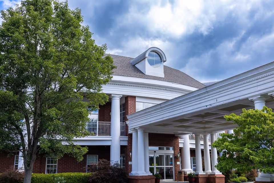 Exterior view of a senior living facility with white columns, a covered entrance, red brick walls, and a large tree in the foreground under a cloudy sky.