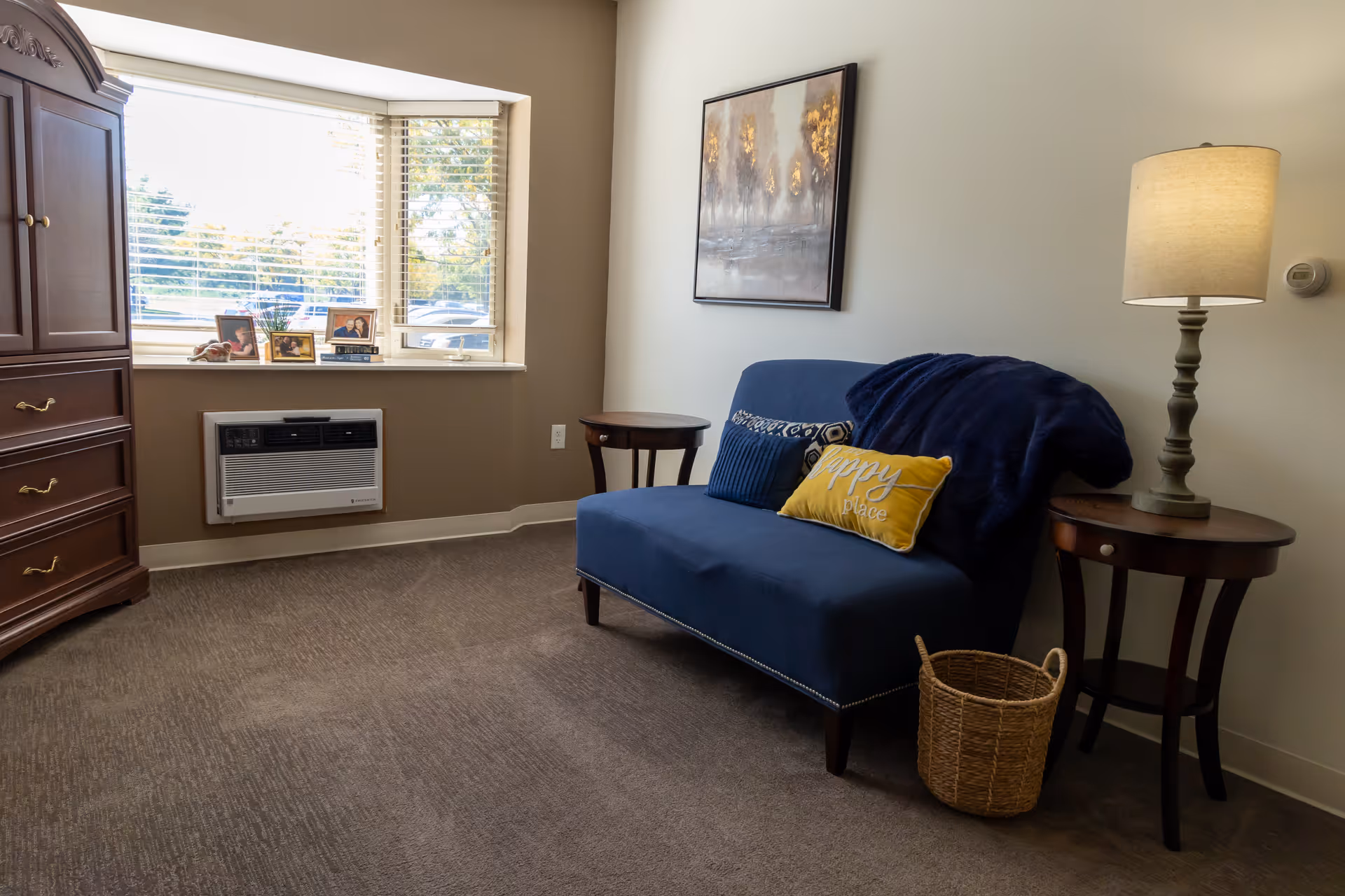 A cozy interior room with a blue loveseat adorned with three decorative pillows and a dark blue throw blanket. On either side of the loveseat are round wooden side tables, one with a lit table lamp. A wicker basket is placed on the floor next to the loveseat. A large wooden dresser is positioned near a window with blinds, which has framed photos on the windowsill. A painting hangs on the wall above the loveseat.