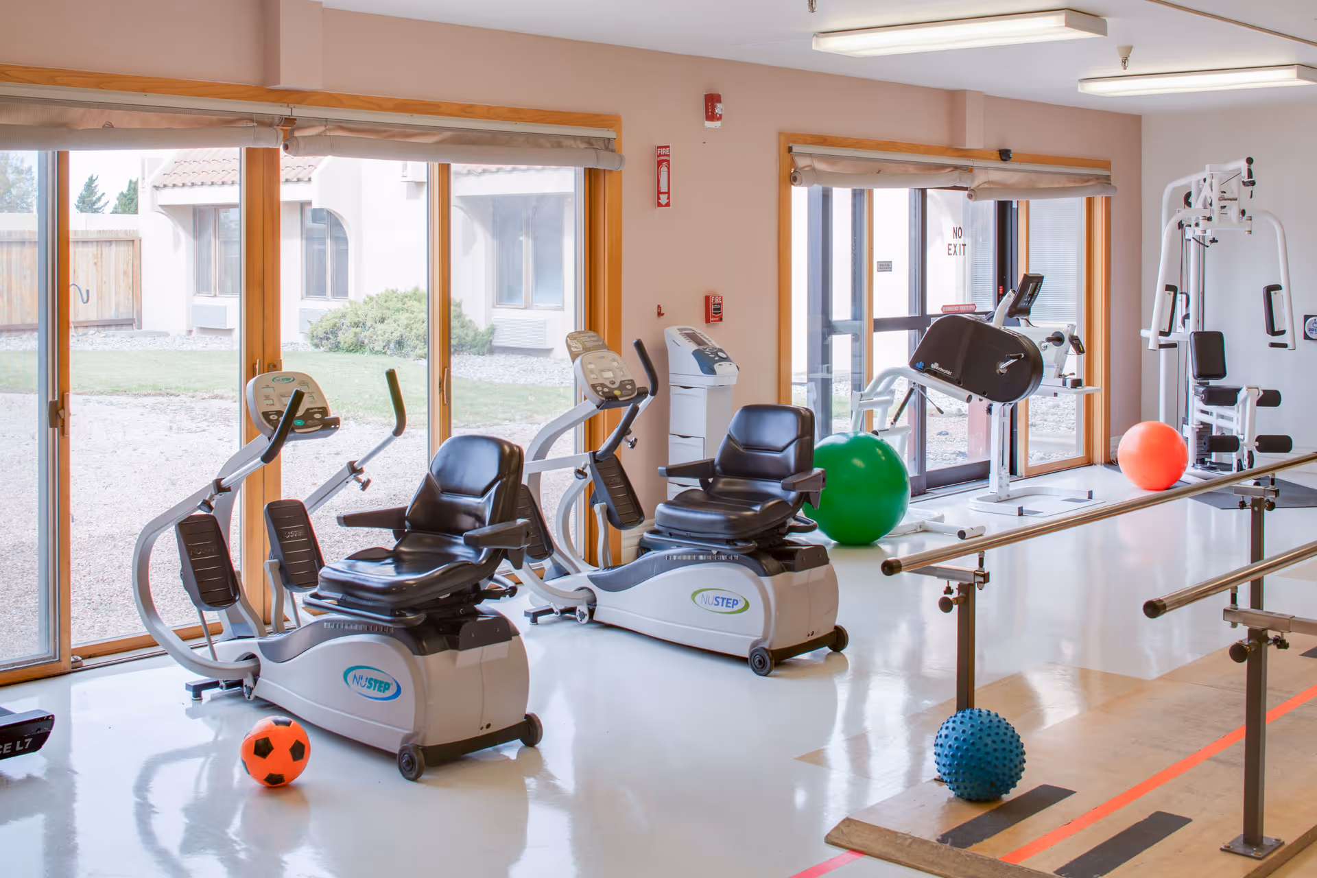Bright therapy/fitness room with recumbent exercise bikes, parallel bars and colorful exercise balls by large sliding windows.