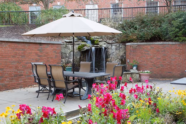 Outdoor patio area with a table and six chairs under a large beige umbrella. Behind the seating area is a stone waterfall feature set against a brick wall. Colorful flowers, including bright pink and yellow blooms, are in the foreground.