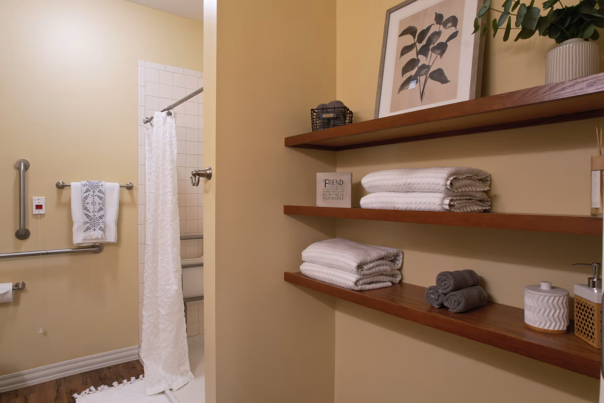 A bathroom area with beige walls featuring a shower with a white textured curtain, grab bars, and a towel rack holding a white towel with a blue pattern. To the right, there are three wooden shelves mounted on the wall holding neatly folded white and gray towels, a framed botanical print, a small plant in a white pot, and decorative containers.
