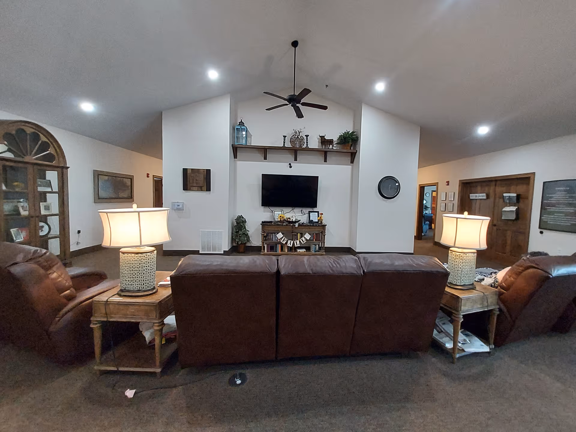 Interior view of a living room area with a brown leather couch and two matching armchairs. Two side tables with lamps flank the couch. A flat-screen TV is mounted on the wall above a wooden console with books and decorative items. The room has a high vaulted ceiling with a ceiling fan and recessed lighting. There are doorways and framed pictures on the walls.