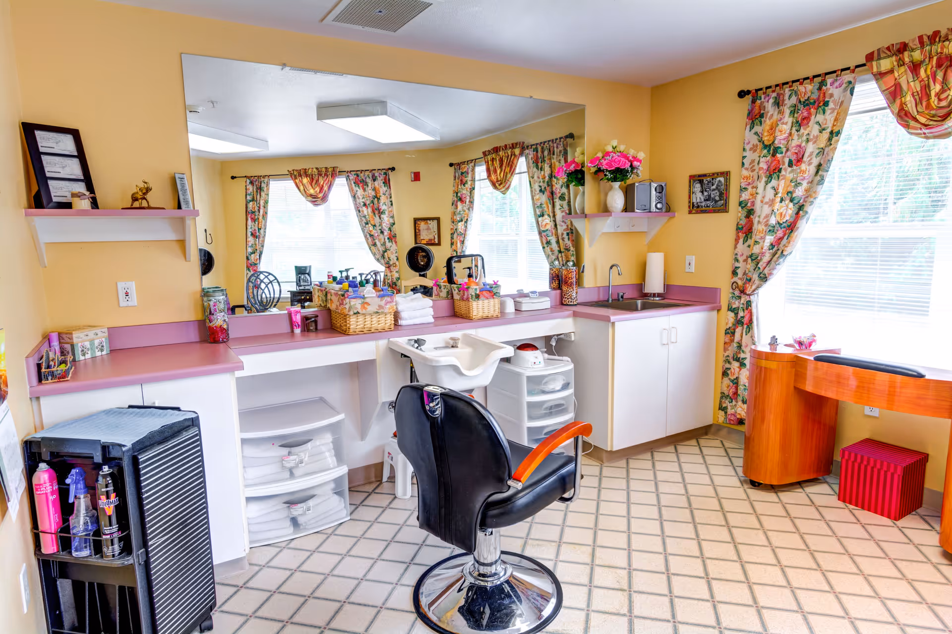 A bright and clean salon area with a black salon chair in front of a white sink and counter. The room has yellow walls, floral curtains on the windows, a large mirror above the counter, and various hair care products and towels neatly arranged. There is also a small wooden desk with a red ottoman underneath near the window.