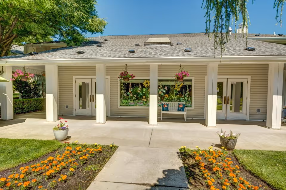 Exterior view of a single-story building with beige siding and a gray roof, featuring a covered porch supported by white columns. The porch has two white doors and a large window decorated with colorful flower and butterfly artwork. There are hanging flower pots and a white bench with decorative pillows under the window. In front of the porch, there are flower beds with orange and yellow flowers and a concrete walkway leading to the entrance. Green trees and a clear blue sky are visible in the background.