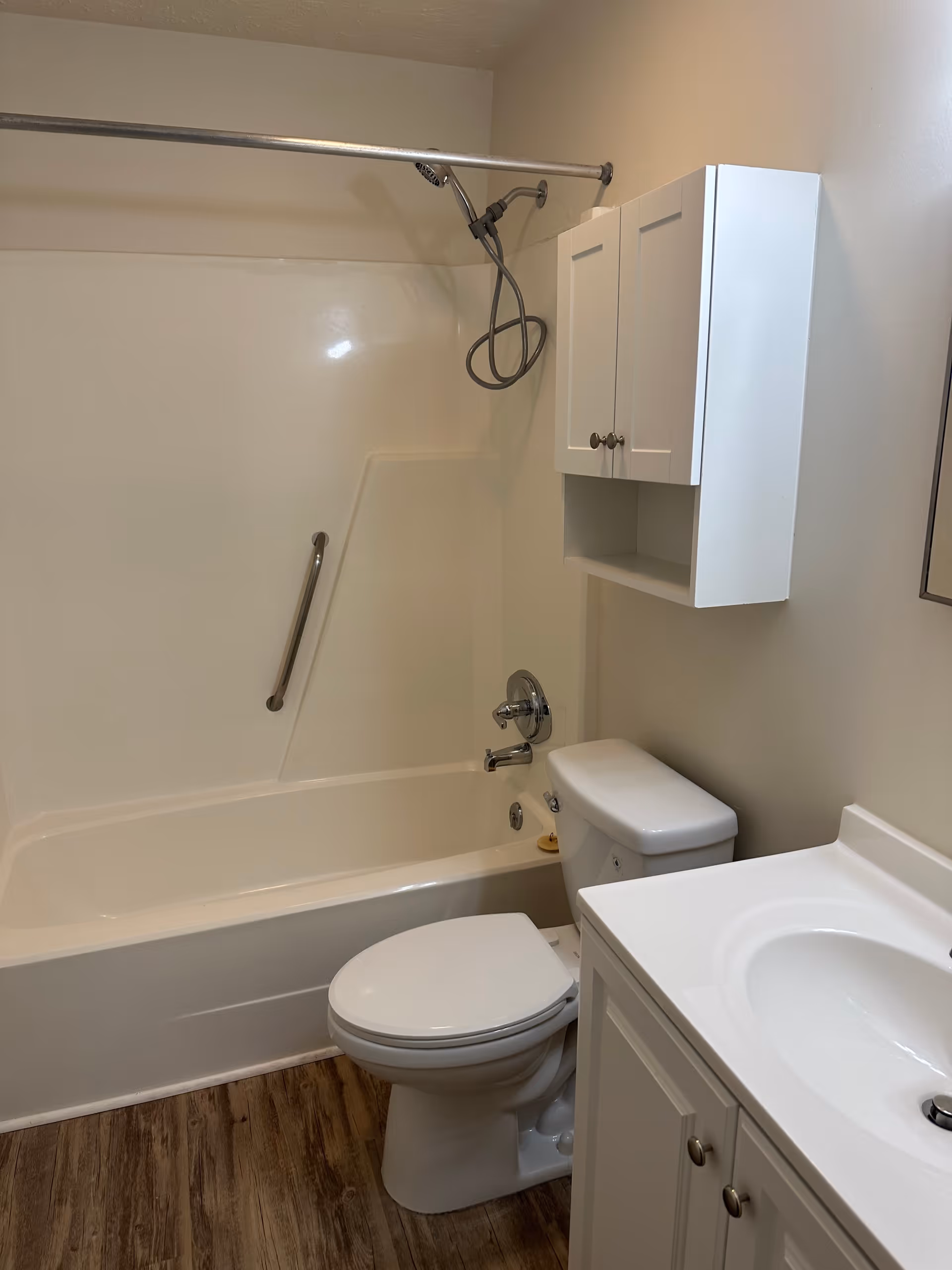 A clean bathroom featuring a white bathtub with a handheld showerhead and a grab bar, a white toilet, a white vanity with a sink, and a white wall-mounted cabinet. The floor has wood-like vinyl flooring.