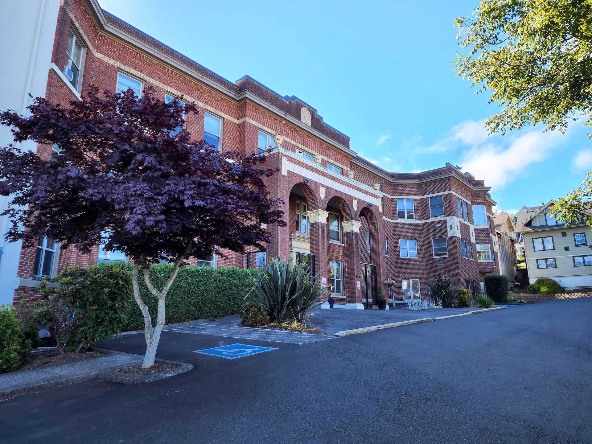 Red-brick multi-story building with arched main entrance, ornamental columns, landscaping and a paved driveway with a visible handicap parking symbol.
