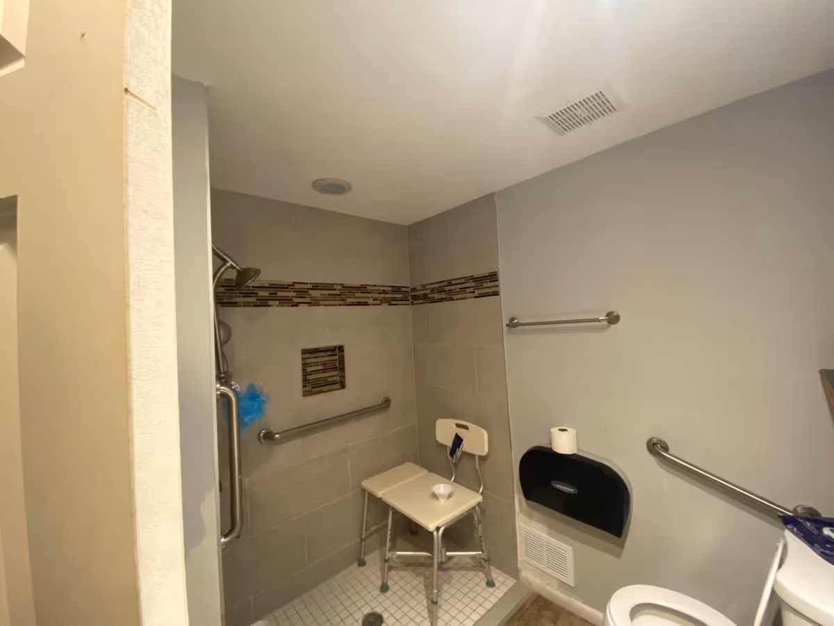 Accessible senior bathroom with a shower chair, grab bars, and tiled walls.