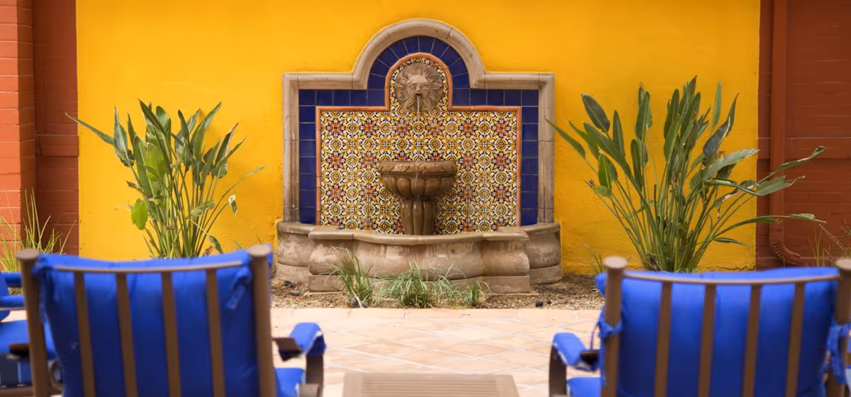 Outdoor seating area with blue cushioned chairs facing a decorative stone fountain mounted on a yellow wall with colorful tile accents, flanked by green plants.