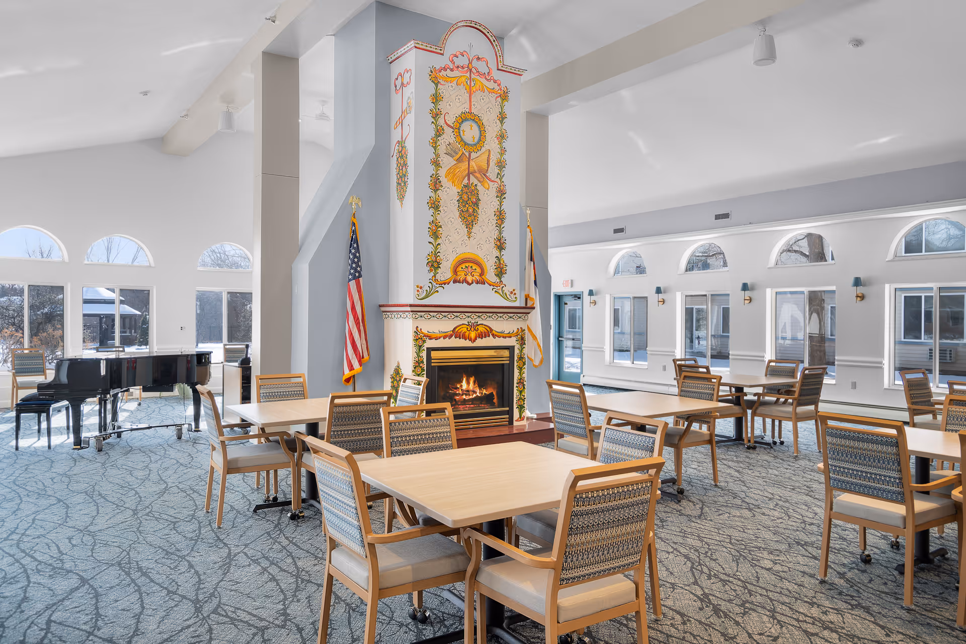 Bright communal dining and activity room with tables and chairs, a decorative fireplace, and a grand piano near arched windows.