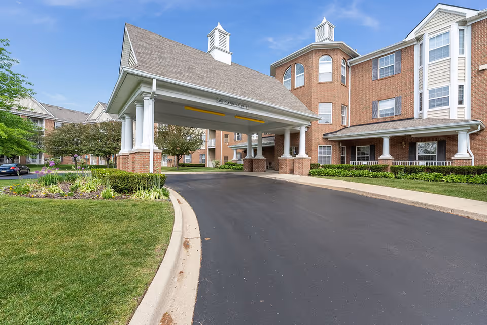 Entrance driveway to a senior living facility with a covered drop-off area supported by white columns. The building is made of red brick with multiple windows and white trim. There is a well-maintained lawn and landscaped garden with flowers and shrubs on the left side of the driveway. The sky is clear and blue.