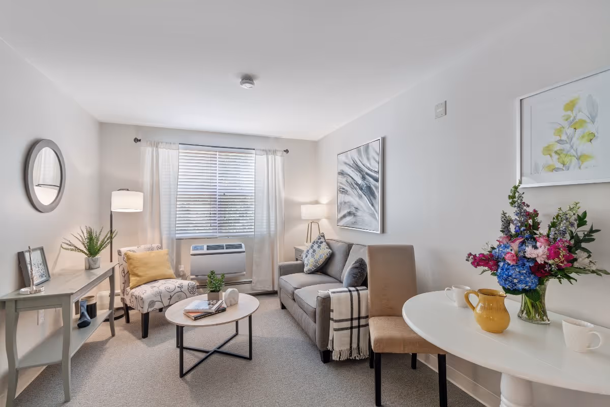 A cozy living room with a gray sofa adorned with a patterned pillow and a throw blanket, a beige upholstered chair, a round white table with a vase of colorful flowers and a yellow pitcher, a small round coffee table with books and a plant, a floor lamp, a console table with a plant and picture frame, and a window with white curtains and blinds letting in natural light.