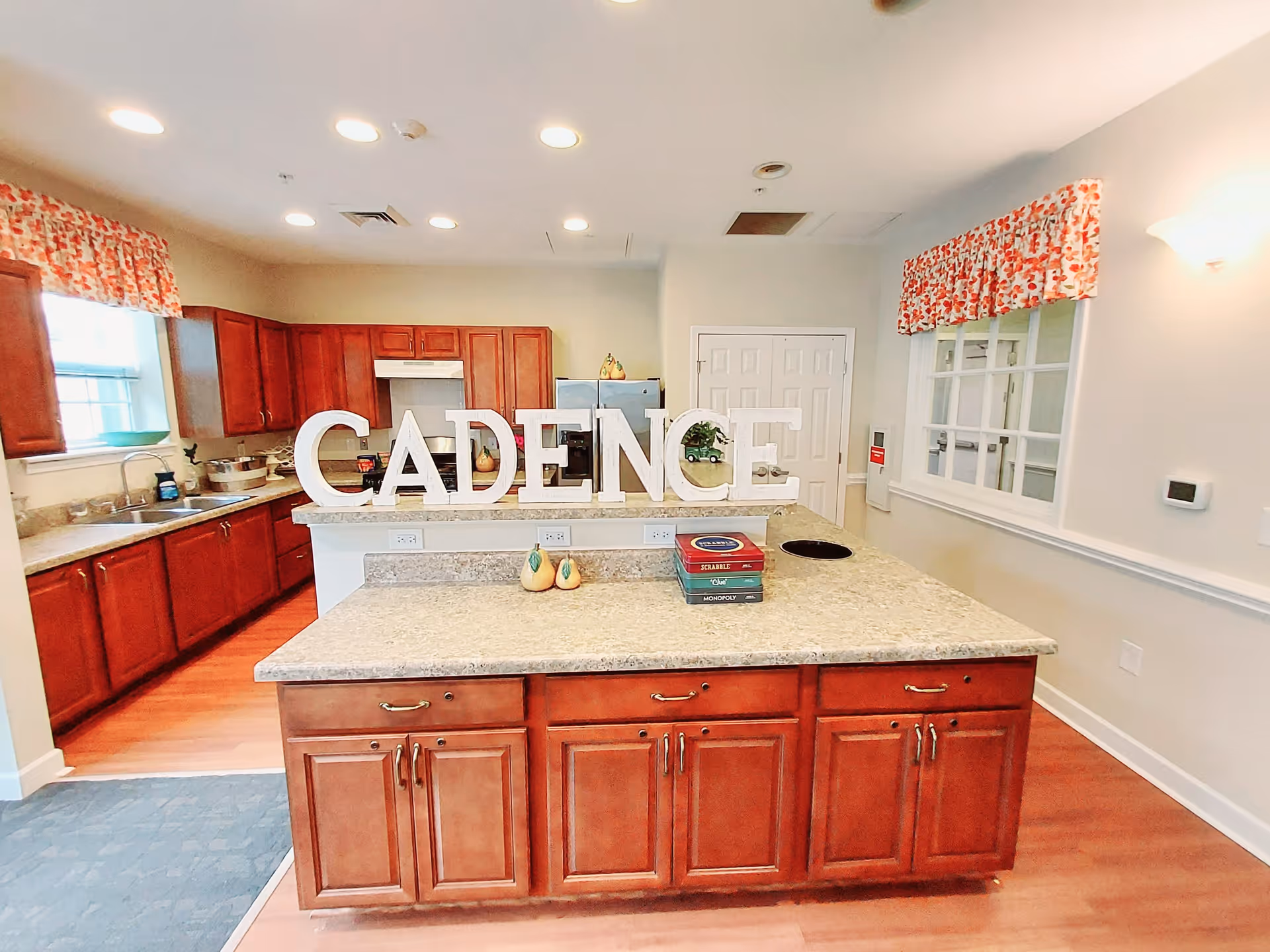 A bright kitchen area with wooden cabinets and a large island countertop in the center. On the island, large white letters spell out 'CADENCE'. The kitchen has a window with floral curtains, a sink, a stove with a hood, and a refrigerator. There are also some decorative items and board games on the island. The walls are light-colored, and the floor is wooden.