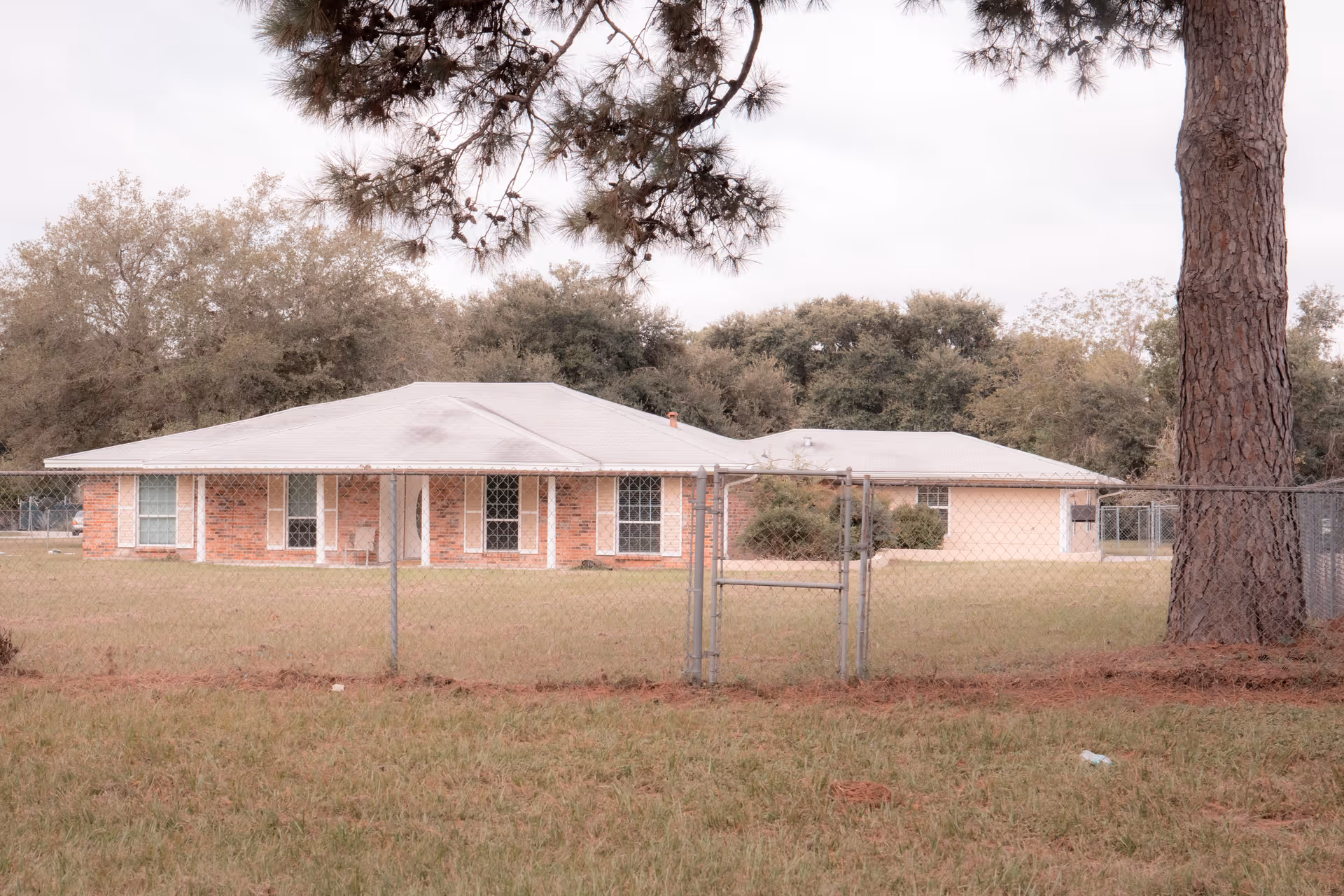 Single-story brick building with a white roof set behind a chain-link fence and large trees in the yard.