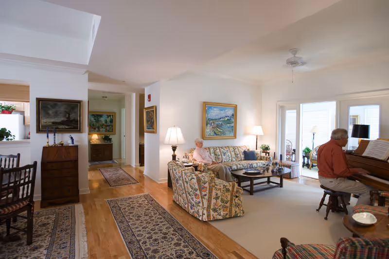 A cozy living room area in a senior living facility with floral patterned sofas and armchairs, a wooden coffee table, and two lamps. An elderly woman is seated on the sofa, and an elderly man is playing a piano. The room has hardwood floors with area rugs and artwork on the walls. There is a dining area visible to the left and a sunroom or enclosed porch with large windows in the background.